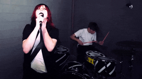 a woman singing into a microphone while a man plays drums in the background