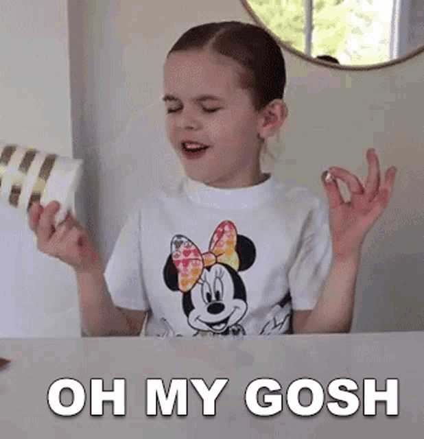 a little girl wearing a minnie mouse shirt is sitting at a table with her hands in the air and says oh my gosh .