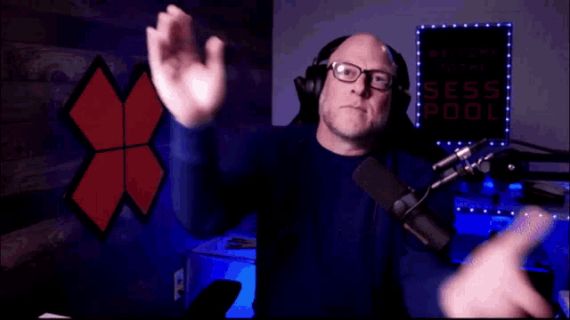 a man wearing headphones and glasses applauds in front of a sign that says welcome to the sess pool