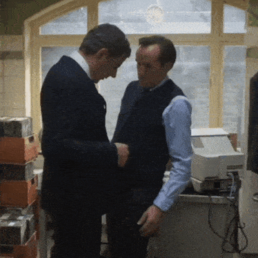 two men standing next to each other in front of a window and a printer