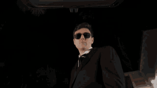 a man wearing sunglasses and a suit is standing in the dark