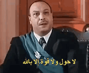 a man in a suit and tie is sitting in a chair with arabic writing on the wall behind him .