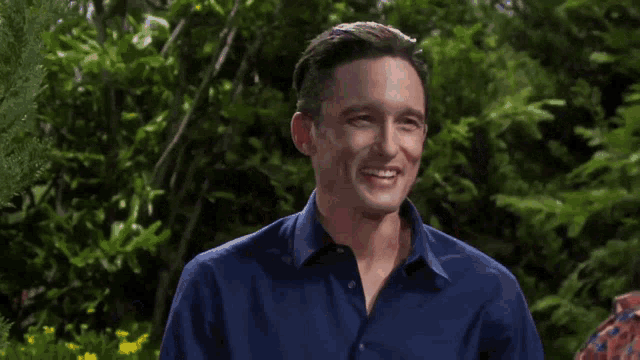 a man in a blue shirt is smiling in front of a forest