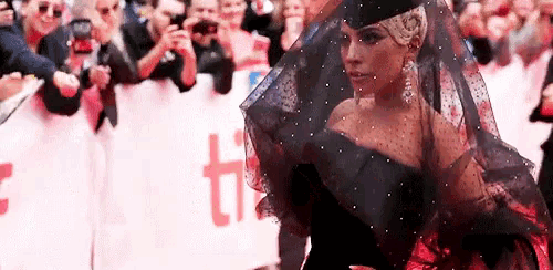 a woman in a black dress and veil is walking on a red carpet in front of a crowd of people .