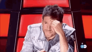 a man in a denim jacket is sitting in a red chair covering his face .