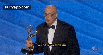 a man in a suit and bow tie is holding a trophy and saying we have work to do .
