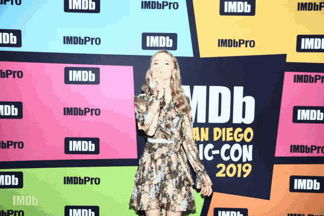 a woman blows a kiss in front of a wall that says imdb an diego ic-con 2019