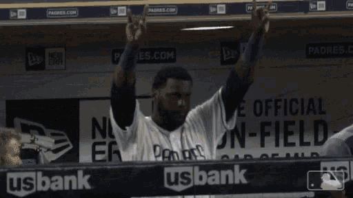 a man in a padres jersey stands in the dugout with his hands in the air