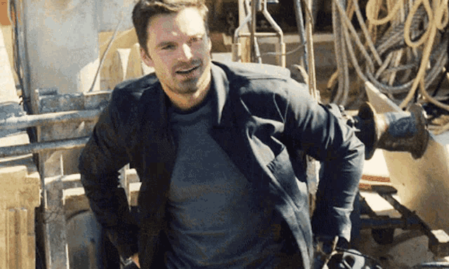 a man wearing a black jacket and a grey shirt is standing in front of a fence