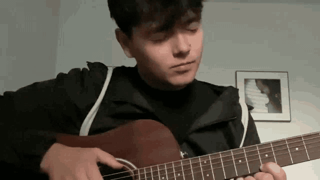 a young man in a black jacket is playing an acoustic guitar in a room .