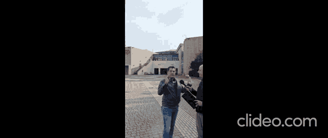 a man is talking into a microphone while another man holds a clipboard in front of a building