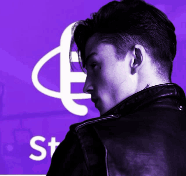 a man in a black leather jacket stands in front of a purple background with the letter s in white letters