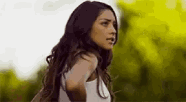 a woman with long hair is wearing a white tank top and standing in front of trees .