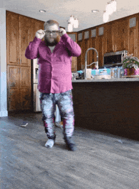 a bearded man in a pink shirt and pants is standing in a kitchen