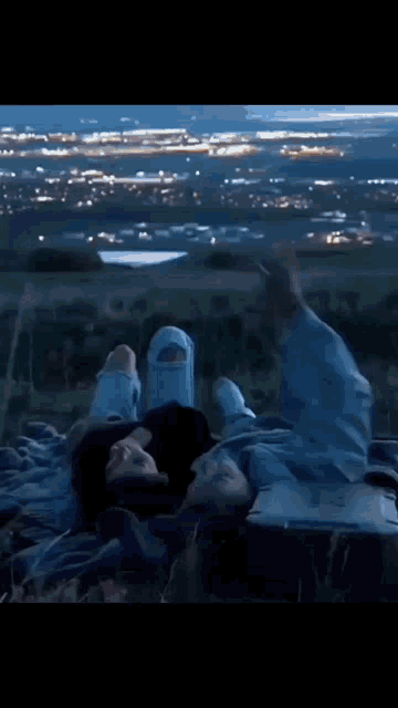 a man and woman are laying on a blanket on top of a hill at night with a city in the background