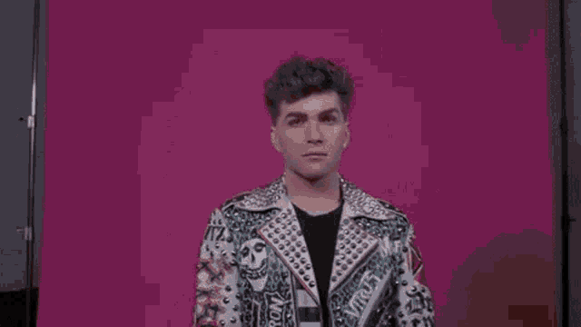 a young man wearing a studded jacket and earrings is standing in front of a pink background .