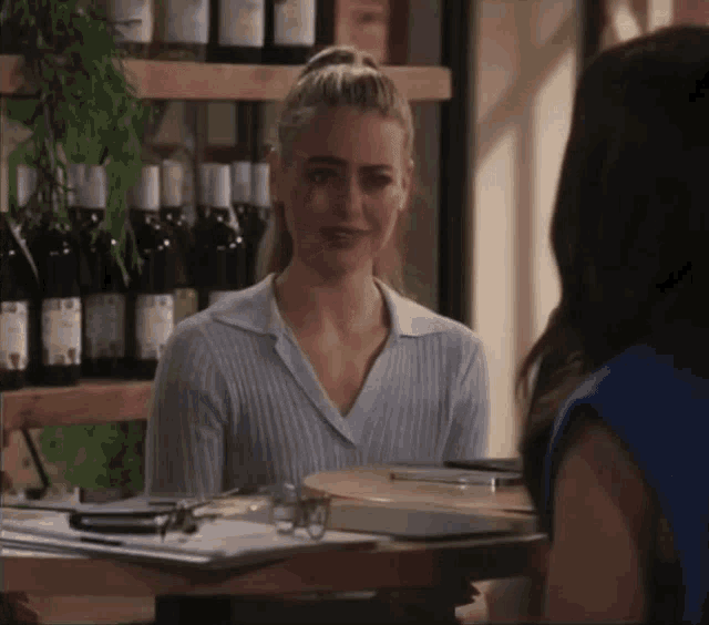 a woman sitting at a table talking to another woman with bottles of wine behind her