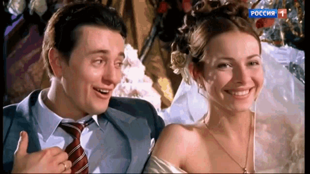 a man in a suit and tie is smiling next to a woman in a wedding dress with a russian tv channel showing