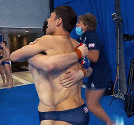 two men hugging each other in front of a blue curtain and a woman wearing a face mask
