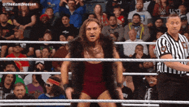a man in a fur coat stands in a wrestling ring while a referee stands behind him