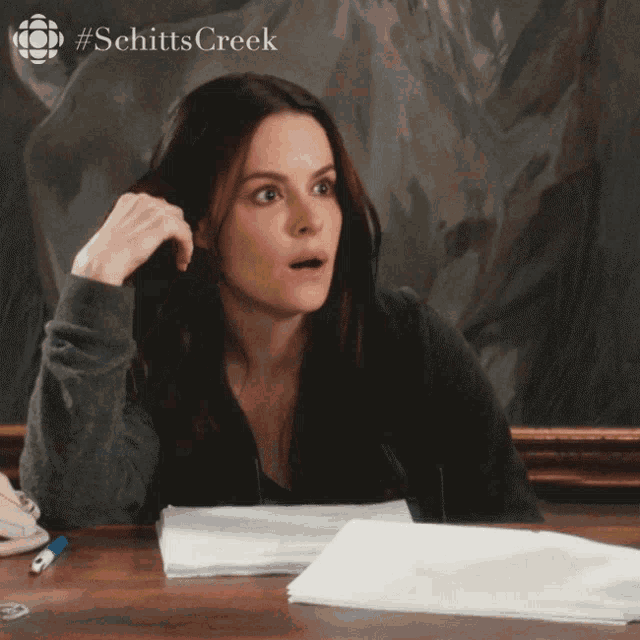 a woman sitting at a desk with a stack of papers and a #schittscreek logo on the bottom