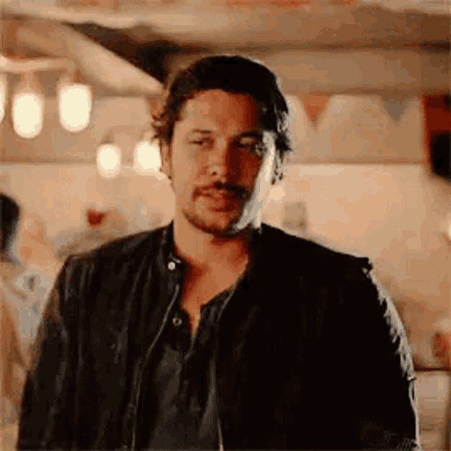 a man with a beard and mustache is wearing a leather jacket and standing in a room .