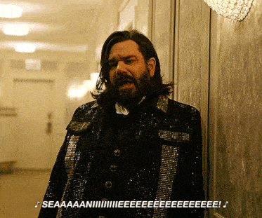a man with long hair and a beard is wearing a sequined jacket and standing in a hallway