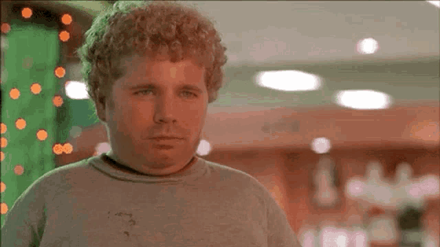 a man with curly hair and a gray shirt is standing in a room with christmas lights in the background .