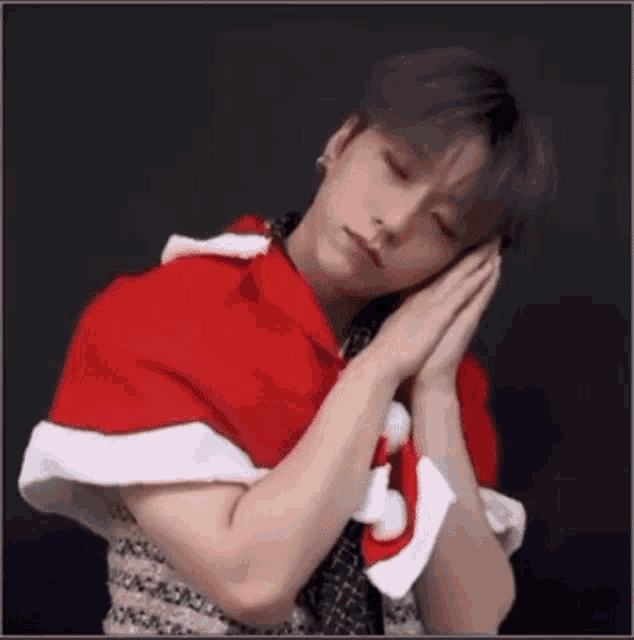 a young man wearing a red and white santa cape is sleeping with his hands on his face .