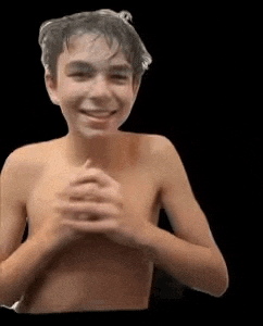 a young boy without a shirt is smiling with his hands folded in front of his chest .
