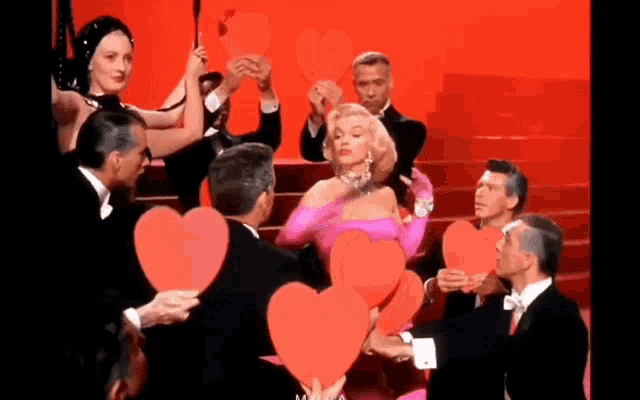 a woman in a pink dress is surrounded by a group of men holding red hearts