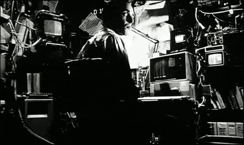 a black and white photo of a man sitting at a desk in a room surrounded by electronics .