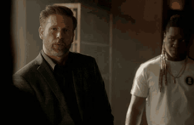 a man in a suit stands next to another man in a white t-shirt
