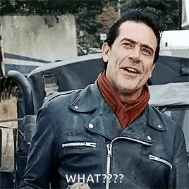 a man wearing a leather jacket and a red scarf is standing in front of a car and talking .