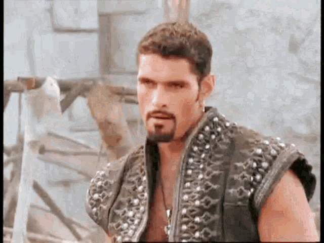 a man with a beard and a studded vest is standing in front of a building and looking at the camera .