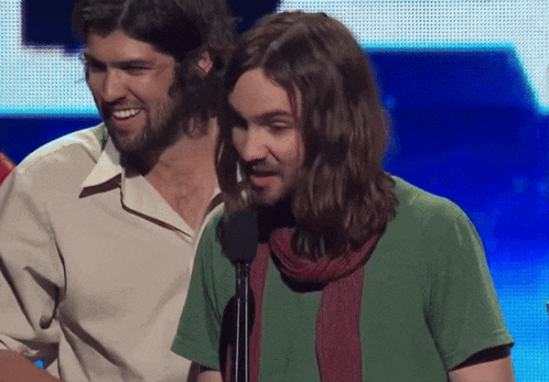 a man with long hair is speaking into a microphone while another man stands behind him