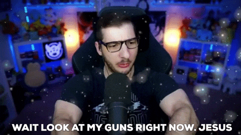a man with glasses is sitting in front of a microphone and says wait look at my guns right now jesus .