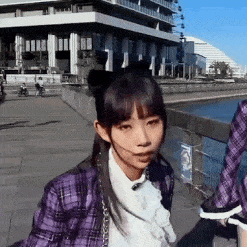 a girl wearing a purple plaid jacket and a white shirt is walking down a sidewalk next to a body of water .