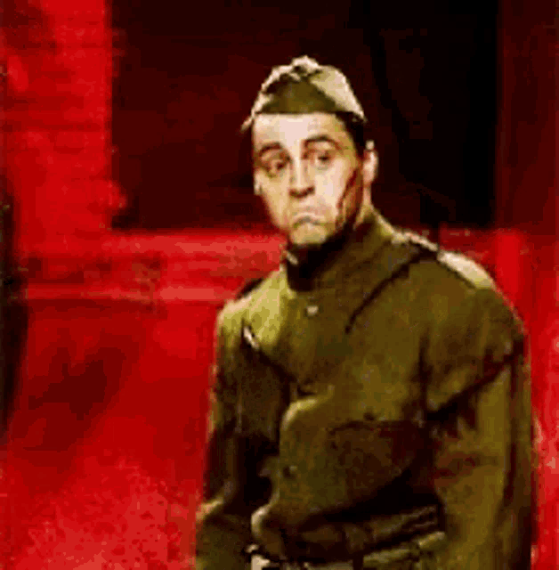 a man in a military uniform is standing in front of a red wall and making a funny face .