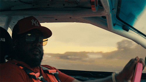 a man wearing a ny hat and sunglasses is driving a car