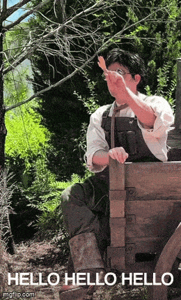 a man is sitting on a wooden bench in the woods and waving .
