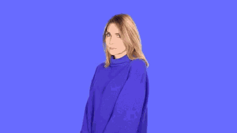a woman in a purple sweater is standing on a blue background and making a funny face .