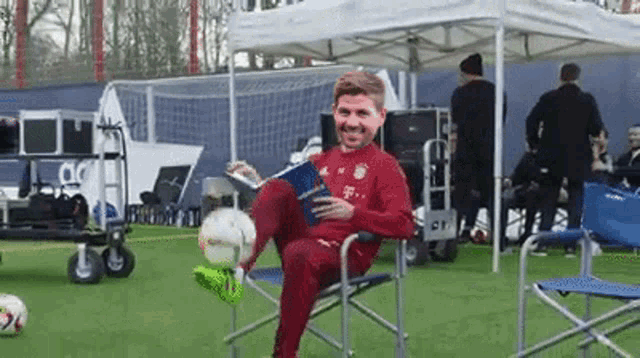 a man is sitting in a chair on a soccer field holding a soccer ball and a book .