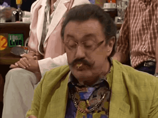 a man with glasses and a mustache is sitting in front of a sign that says live