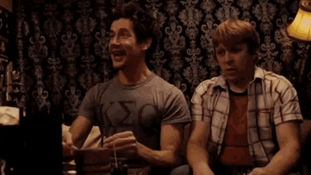two men are sitting at a table and laughing while one of them is holding a cell phone .