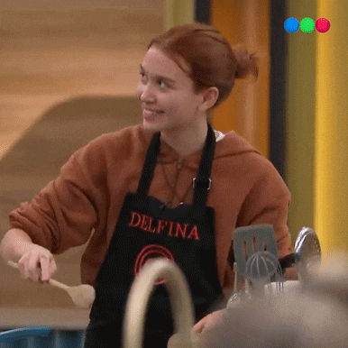 a woman wearing an apron that says delfina is holding a wooden spoon and smiling .