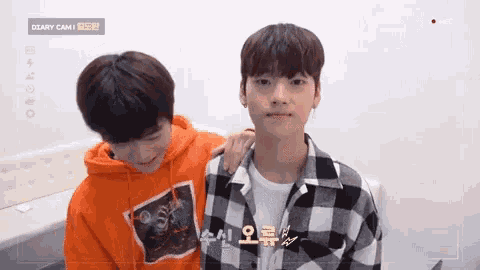 two young men are sitting next to each other and one of them is wearing an orange hoodie and a plaid shirt .
