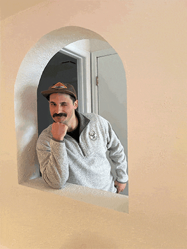 a man wearing a hat and a grey sweater looks out of an arched window