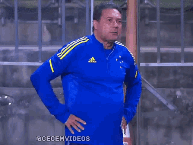 a man wearing a blue adidas sweater stands with his hands on his hips