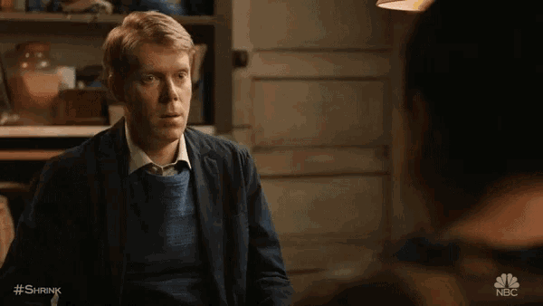 a man in a suit and blue sweater is talking to another man with #shrink written on the bottom of the screen